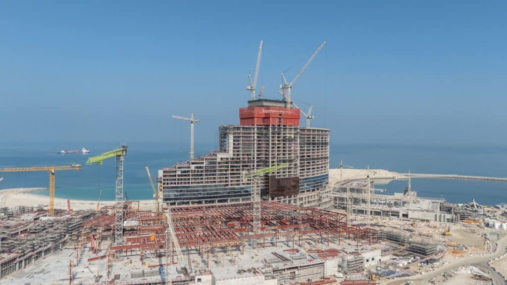 A large, partially constructed Wynn Al Marjan Island building with multiple cranes on a coastal site near the sea.
