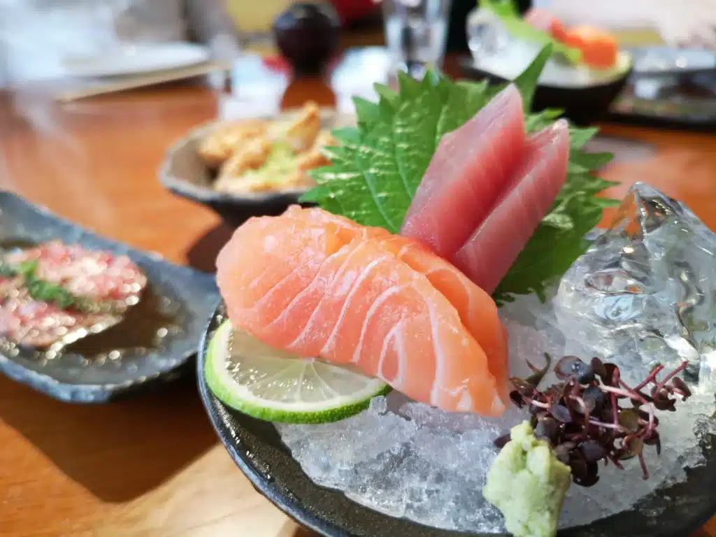 A plate of sashimi featuring salmon and tuna slices on ice, garnished with shiso leaves, lime, and wasabi.