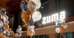 Balloons in blue, gold, and white decorate an indoor space with a bright "zuma" sign on a wall.