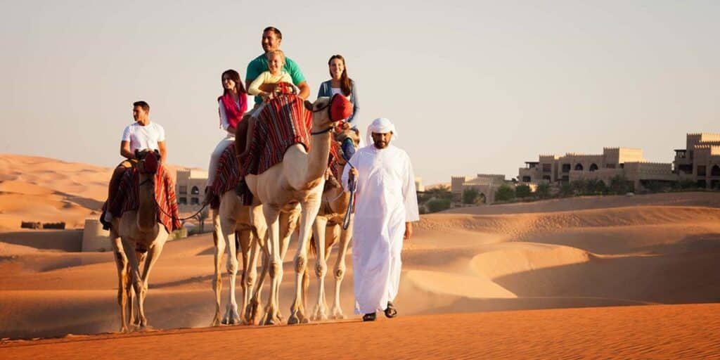 People riding camels led by a guide in traditional attire, with a backdrop of desert sand dunes and distant buildings.