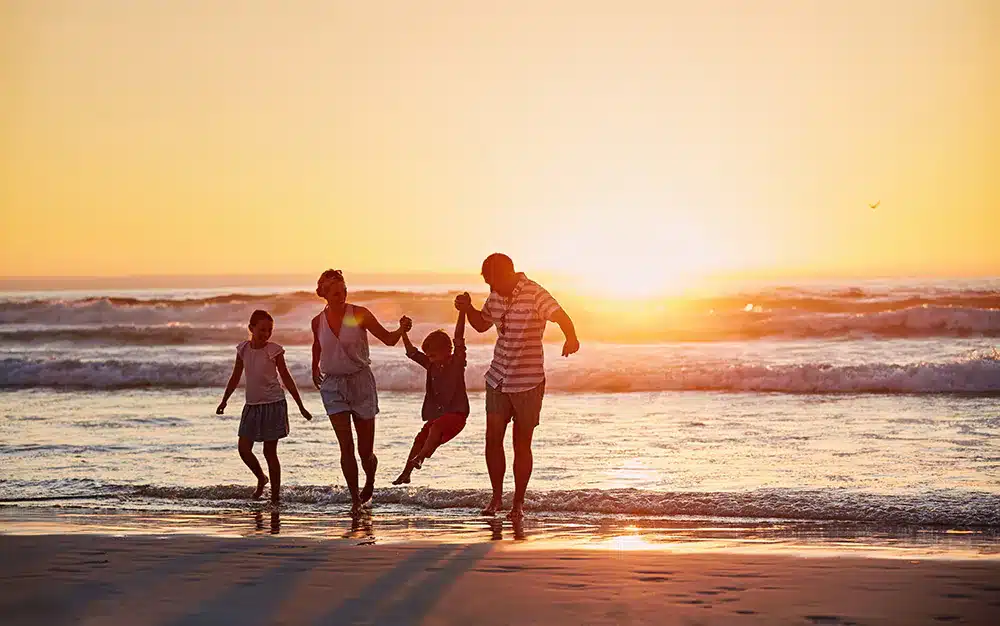 A family of four holds hands while walking along a beach at sunset.