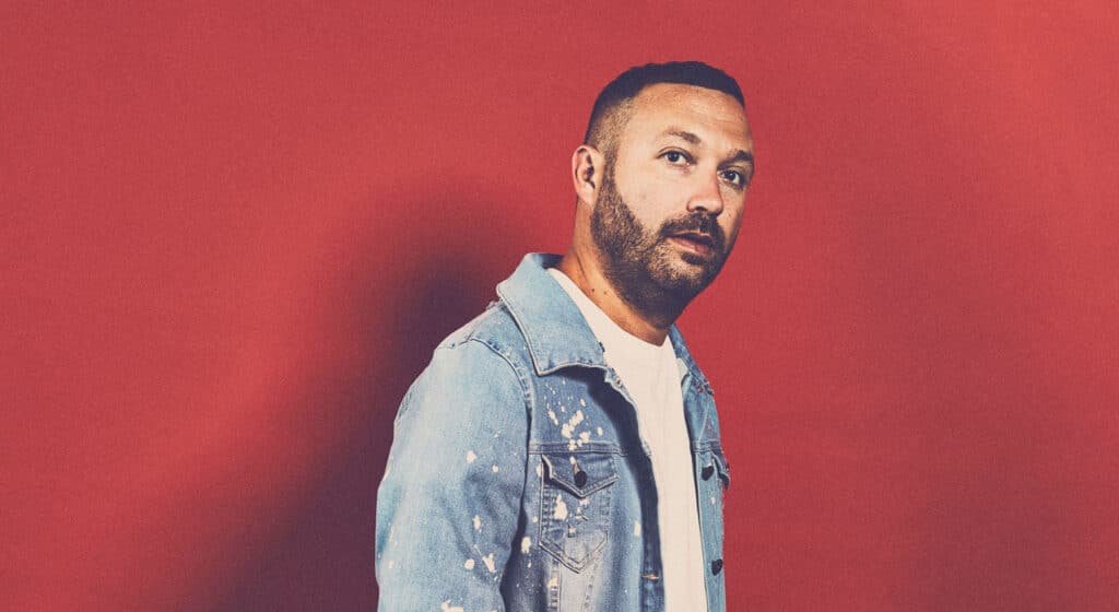 A man with a beard and denim jacket stands against a red background.