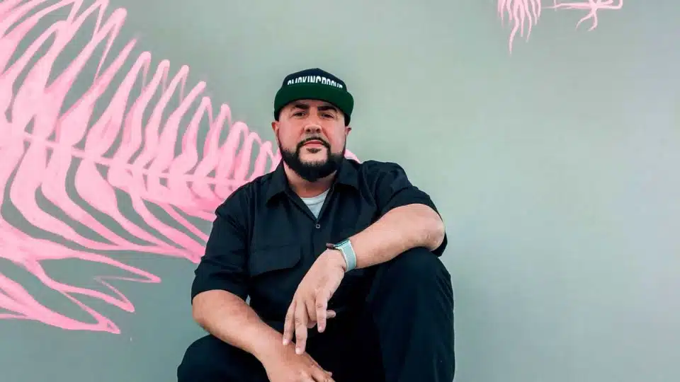 Smokingroove in a black shirt and cap sitting in front of a wall with pink abstract art