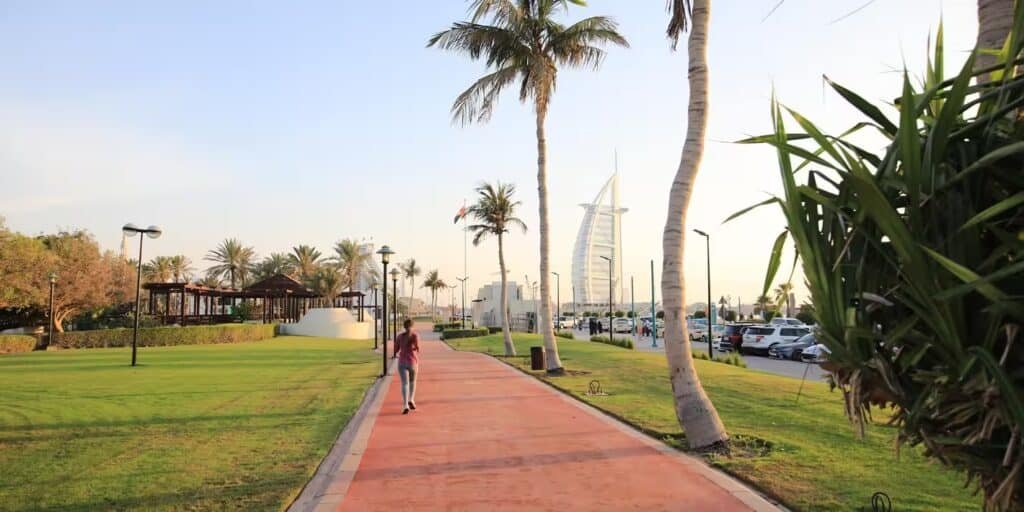 Person walking on a palm tree-lined path with the Burj Al Arab visible in the background.