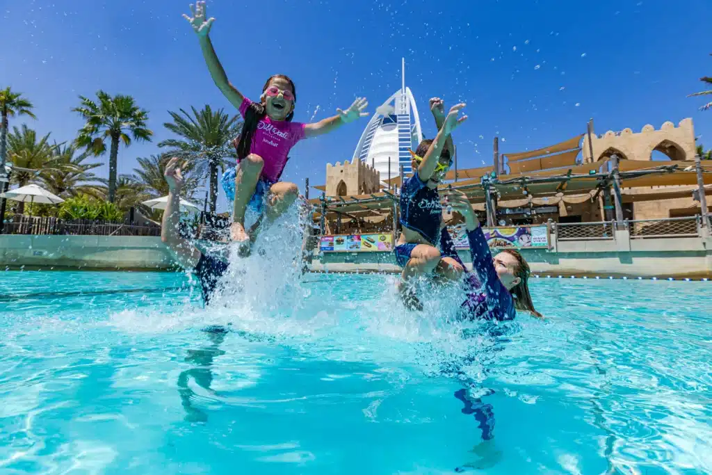 Children joyfully playing and splashing in a sunny Wild Wadi Waterpark with a distinctive sail-shaped building in the background.