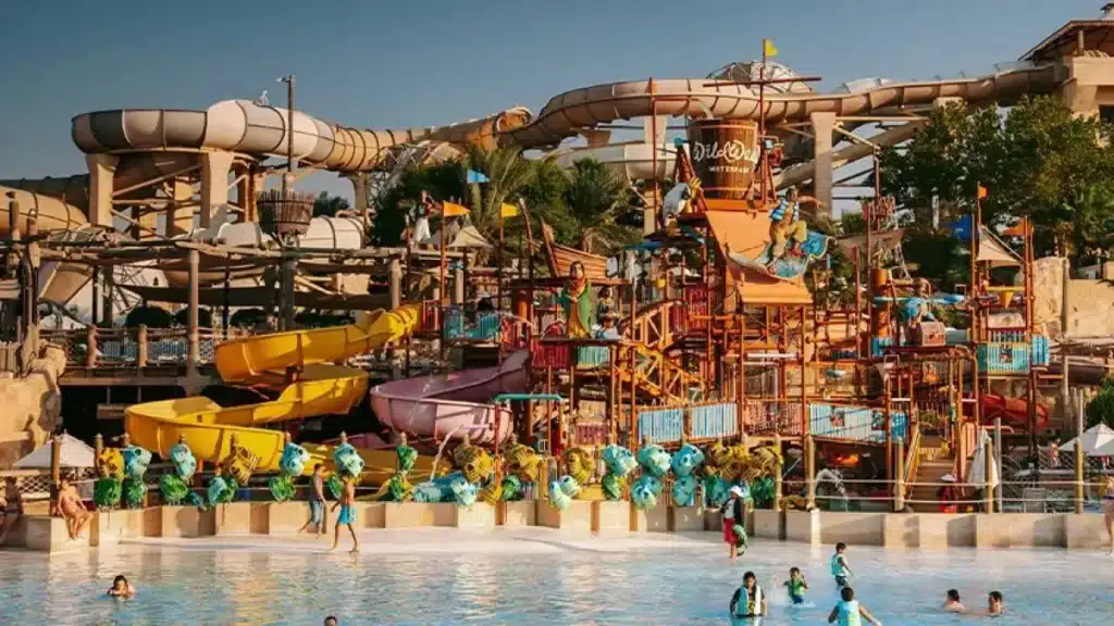 A bustling water park with numerous winding slides, colorful play structures, and people enjoying the wave pool.