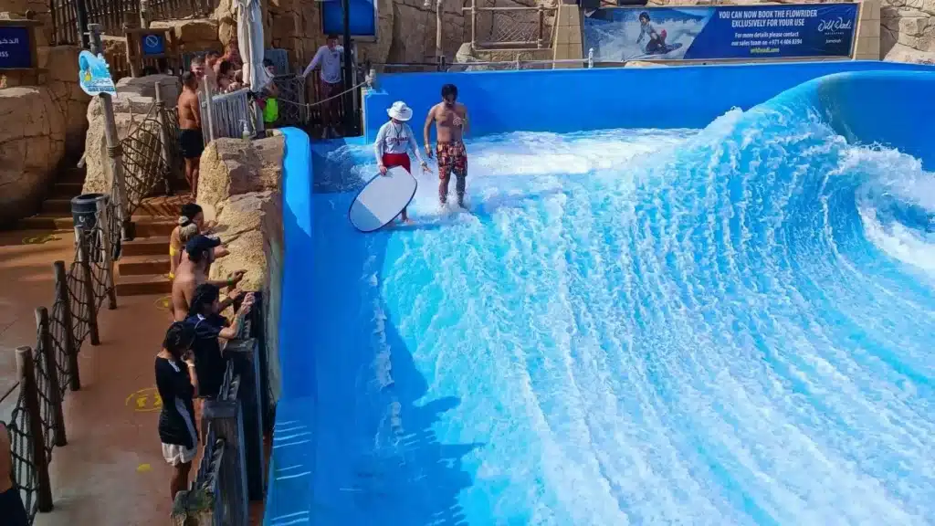 Two people stand on a blue artificial wave machine, one holding a surfboard, with onlookers watching from the side.