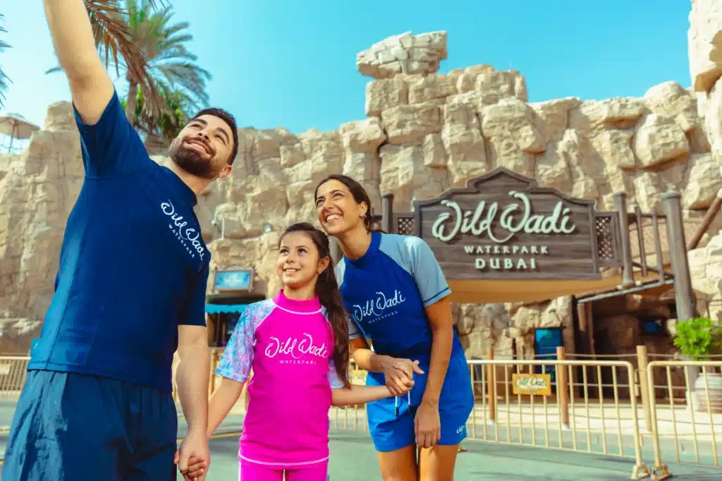 A family of three in colorful clothing smiles and takes a selfie in front of the Wild Wadi Waterpark entrance in Dubai.