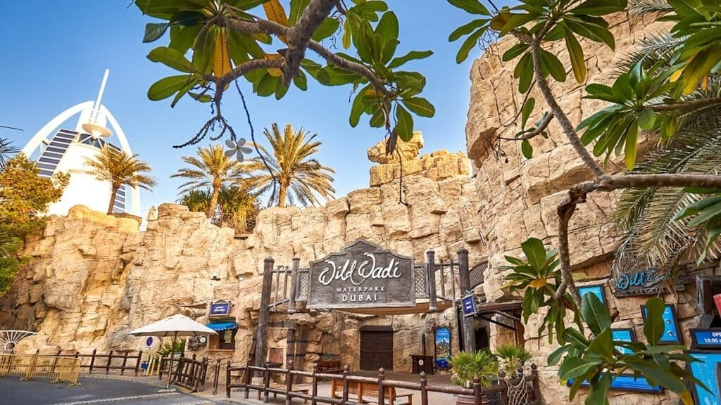 Entrance to Wild Wadi Waterpark in Dubai, featuring rocky structures and palm trees, with the Burj Al Arab visible in the background.