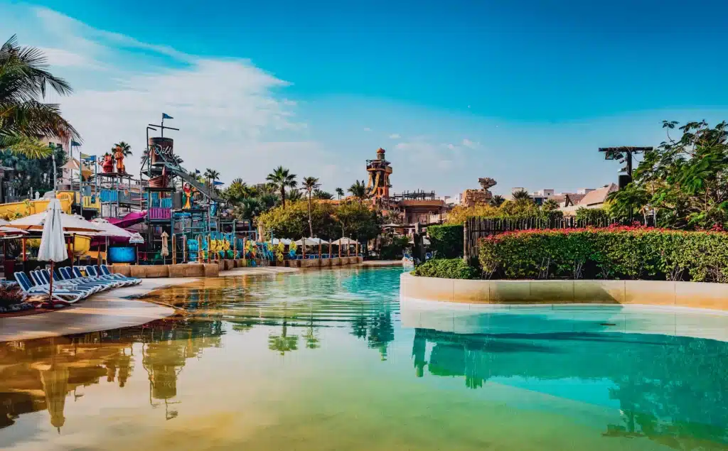 A tranquil water park scene with a clear pool, colorful slides, and lounge chairs under umbrellas, surrounded by lush greenery.