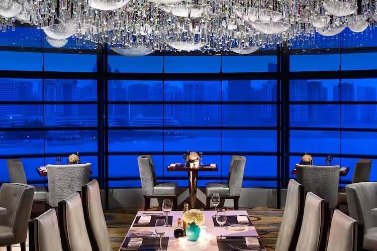 Elegant restaurant interior with modern decor, featuring soft lighting and a view of a city skyline through large windows.