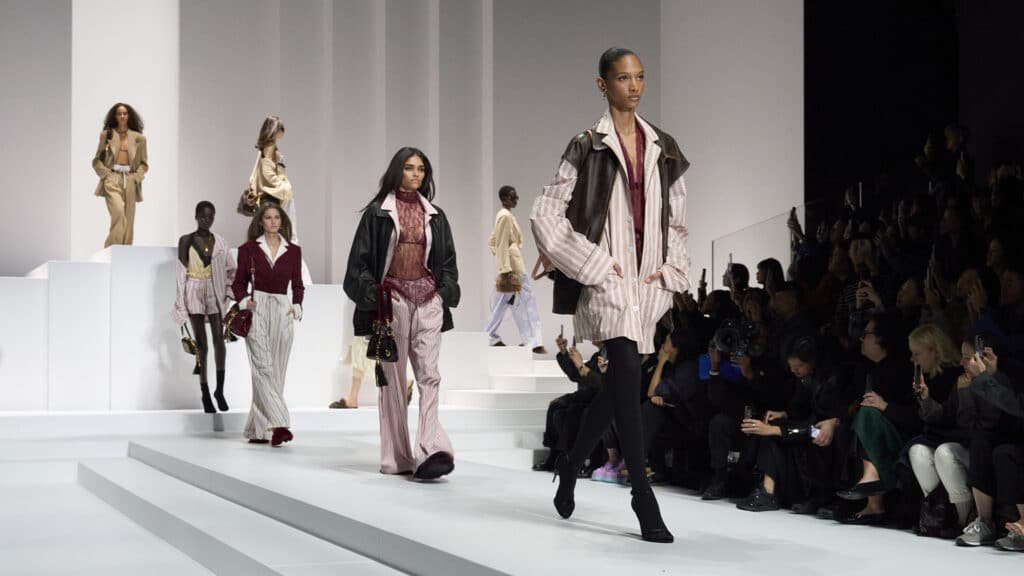 Models walk down a fashion runway wearing elegant outfits in a contemporary show setting.