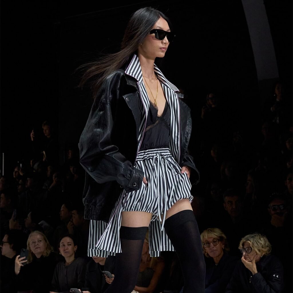 Model walks runway in black leather jacket, striped shorts, and sunglasses, set against a dark crowd-filled backdrop.