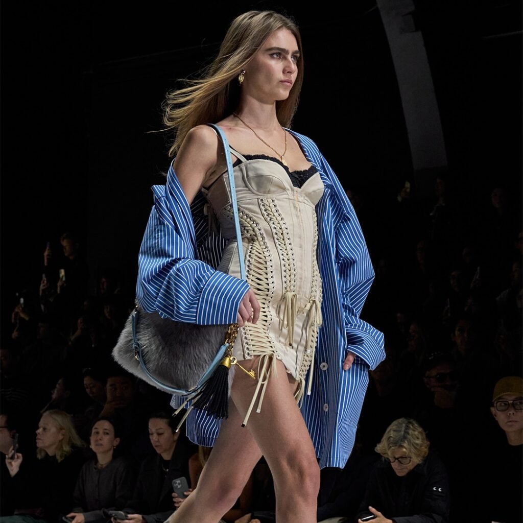 A model walks the runway wearing a striped blue shirt over a corset-style beige top, with a fur-accented handbag.