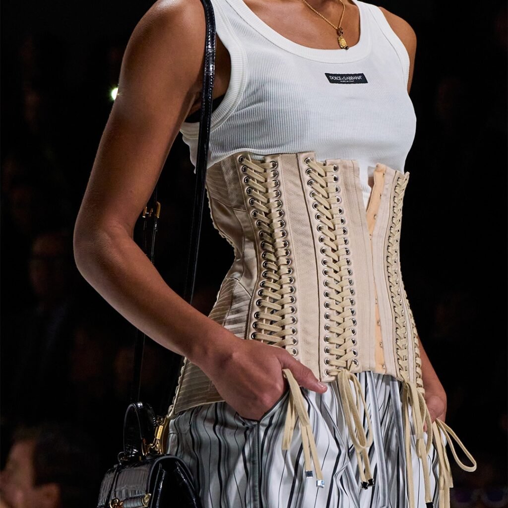 A model wearing a white Dolce & Gabbana tank top with a beige corset featuring lace-up details and striped pants, holding a black bag.