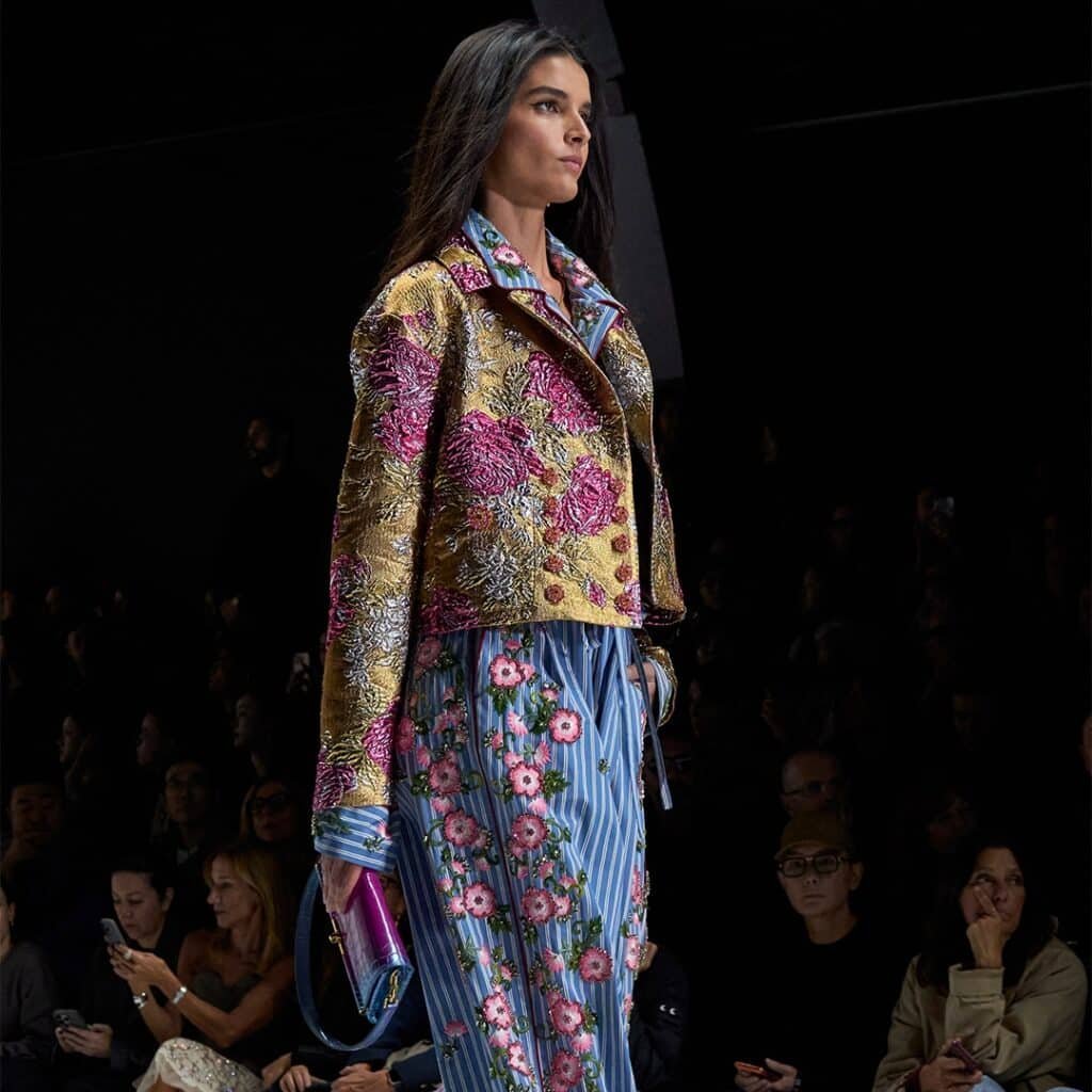 A model walks the runway wearing a colourful Dolce & Gabbana floral jacket and striped floral pants, holding a purple clutch, with an audience in the background.