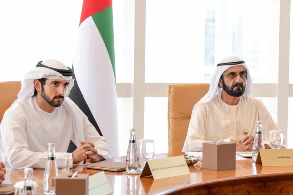 Sheikh Hamdan bin Mohammed bin Rashid Al Maktoum and Sheikh Mohammed bin Rashid Al Maktoum in white traditional attire, sit at a conference table with a UAE flag in the background.