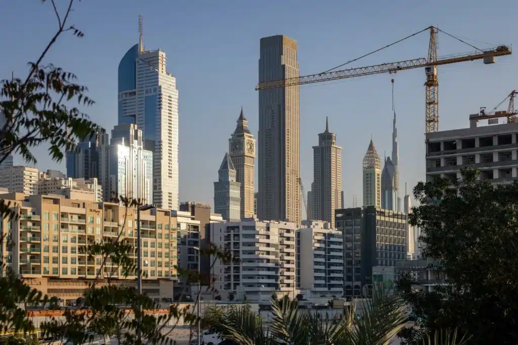 City skyline with modern Dubai Home Prices skyscrapers and a construction crane in the foreground, trees partially framing the view.