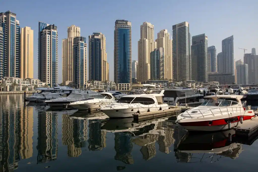 Yachts docked at a marina with a backdrop of tall, modern skyscrapers under a clear sky.