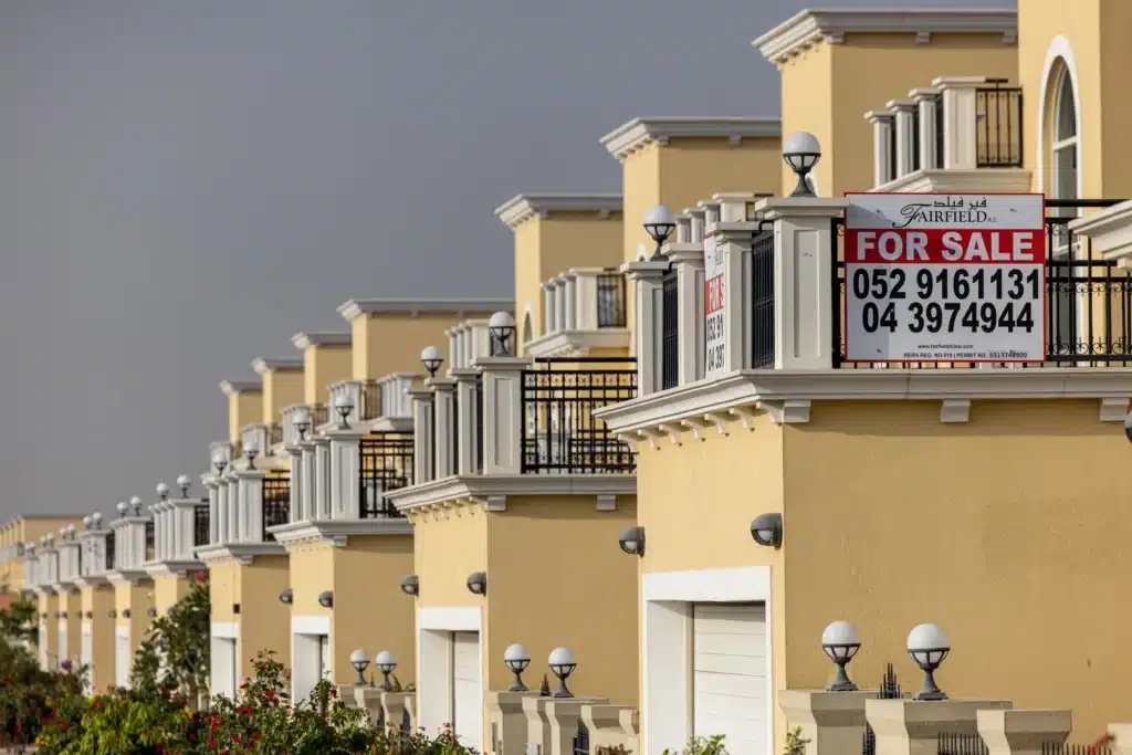 A row of beige townhouses with balconies and a "For Sale" sign featuring contact numbers.
