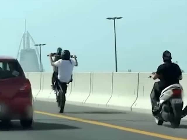 Two motorcyclists perform wheelies on a highway, with skyscrapers visible in the background.