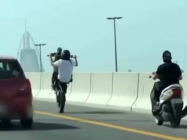 Two motorcyclists perform wheelies on a highway, with skyscrapers visible in the background.