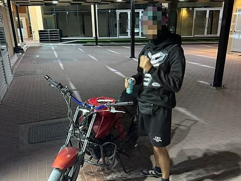 A person wearing a hoodie and shorts stands near a red motorcycle holding a beverage can, with their face pixelated.