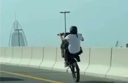 A person wearing a helmet performs a wheelie on a motorcycle on a bridge, with a tall, pointed building visible in the background.