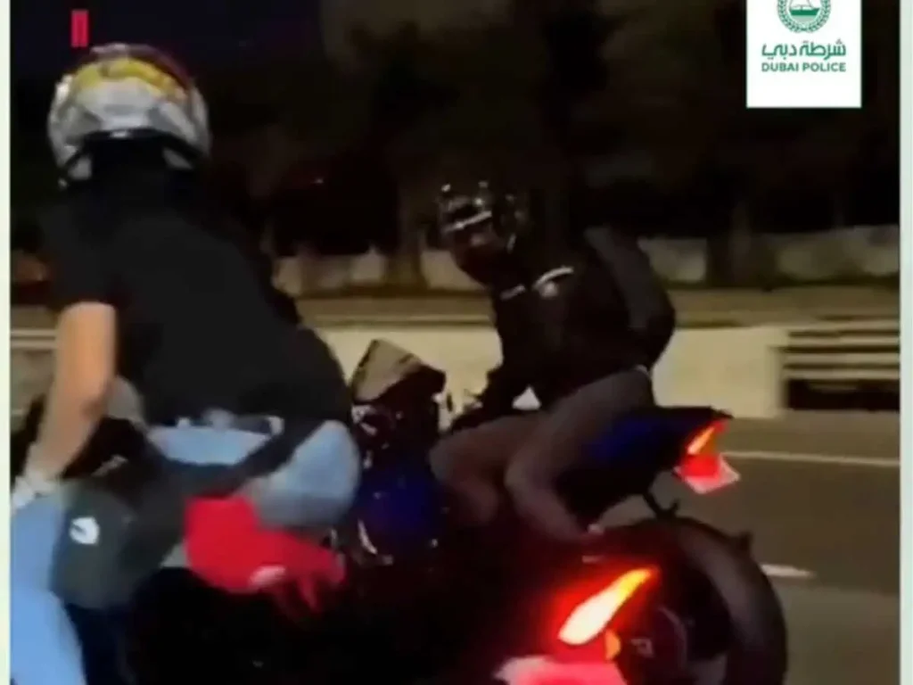 Two motorcyclists in helmets riding at night on a street, with Dubai Police logo in the corner.