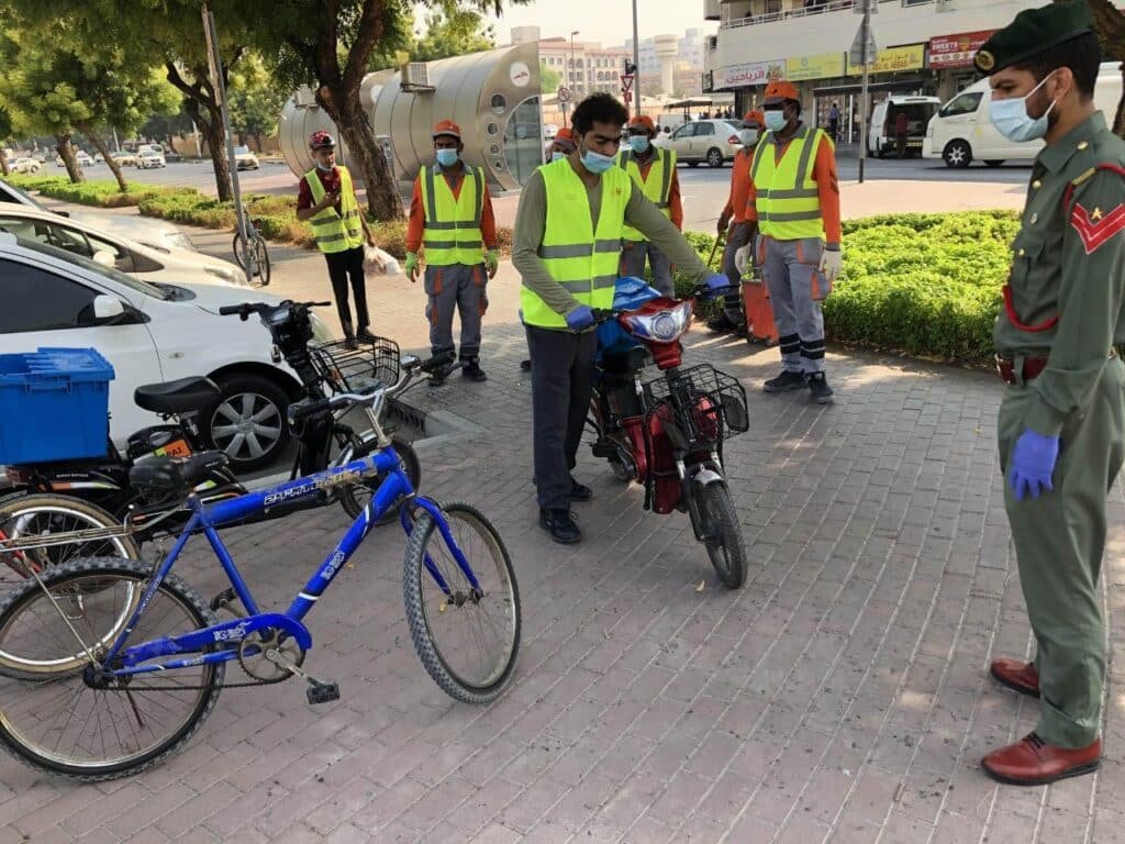 Dubai Police Seize Bikes: Group of workers in reflective vests and masks standing with bicycles and an electric scooter on a paved area near parked cars.