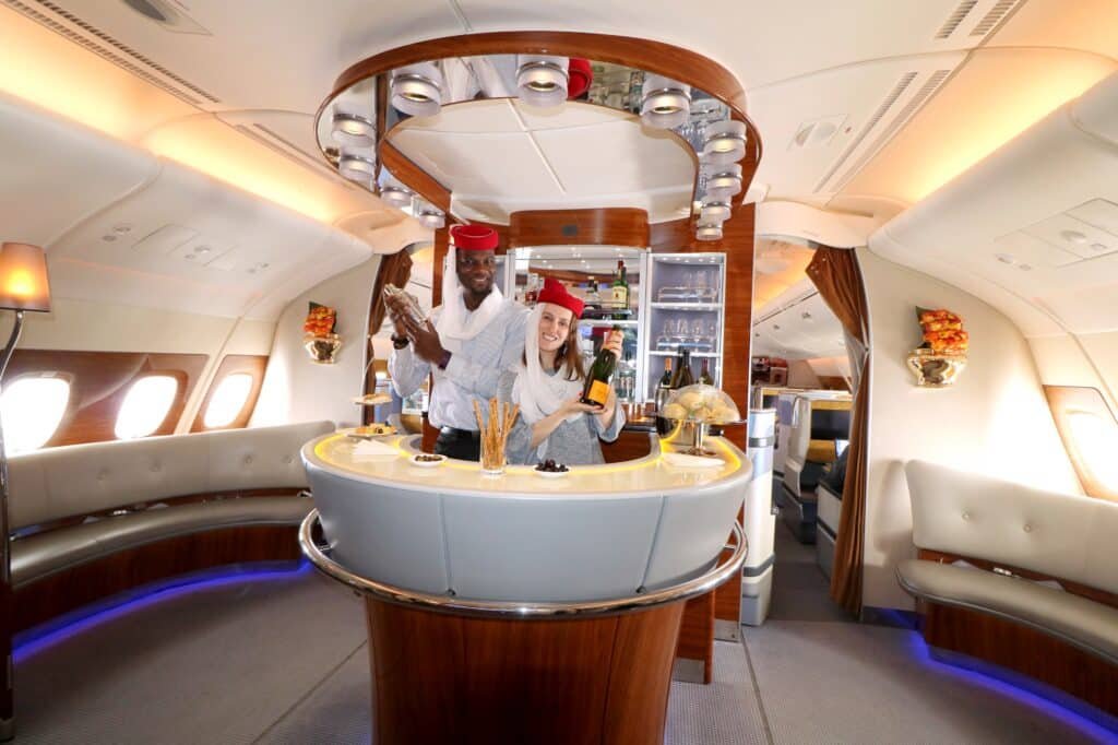 Two flight attendants in uniform serve drinks at a luxurious airplane bar with elegant decor and seating.