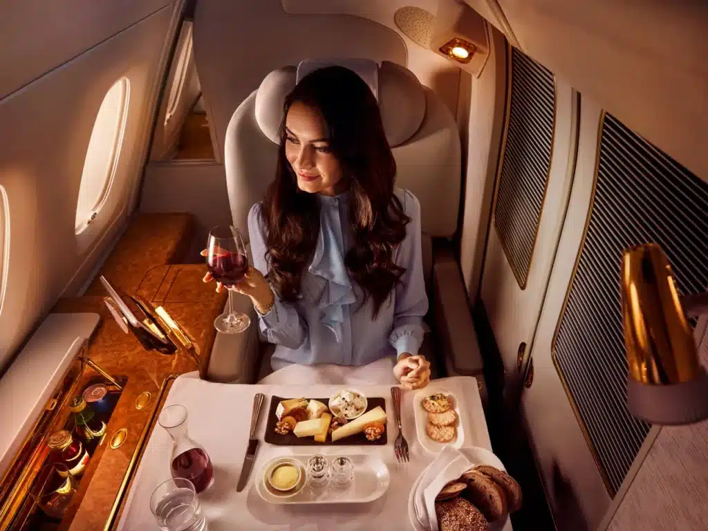 A woman enjoys a meal while seated in a luxurious Emirates First Class A380 airplane cabin, holding a glass of red wine.
