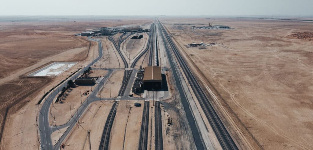 Aerial view of a vast desert landscape with a Etihad Rail Full List Of Stations yard featuring multiple tracks, buildings, and surrounding barren terrain.