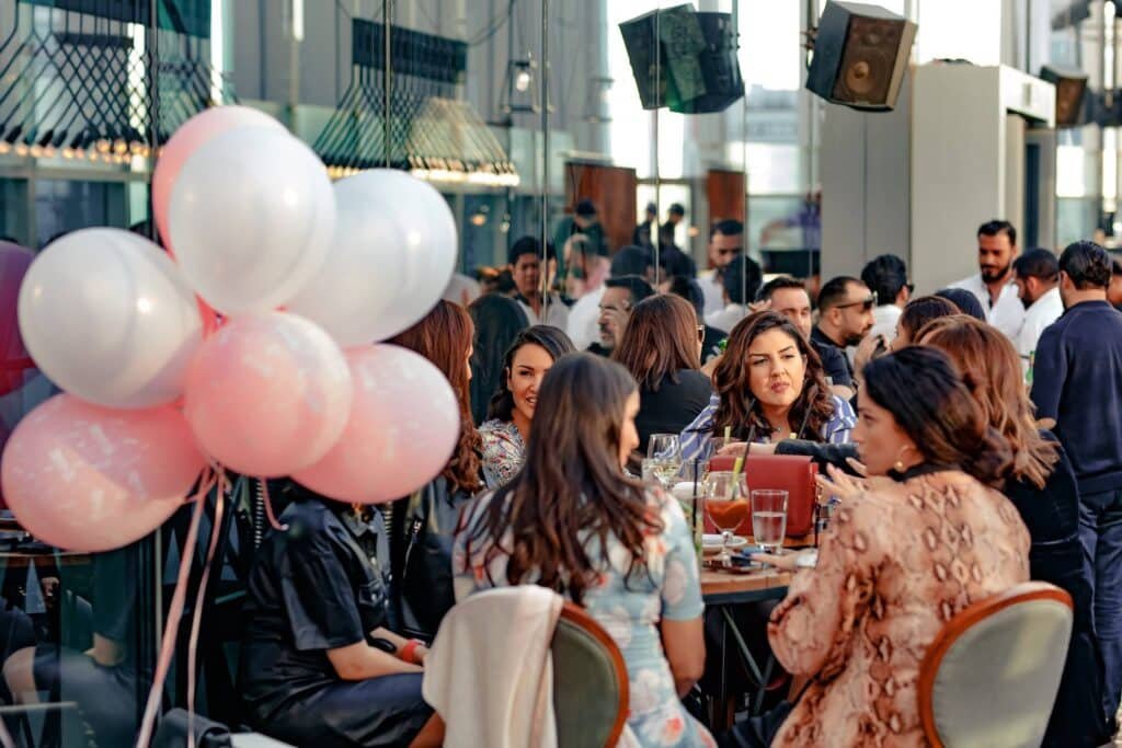Group of people seated at a table in a lively outdoor gathering, with pink and white balloons nearby.