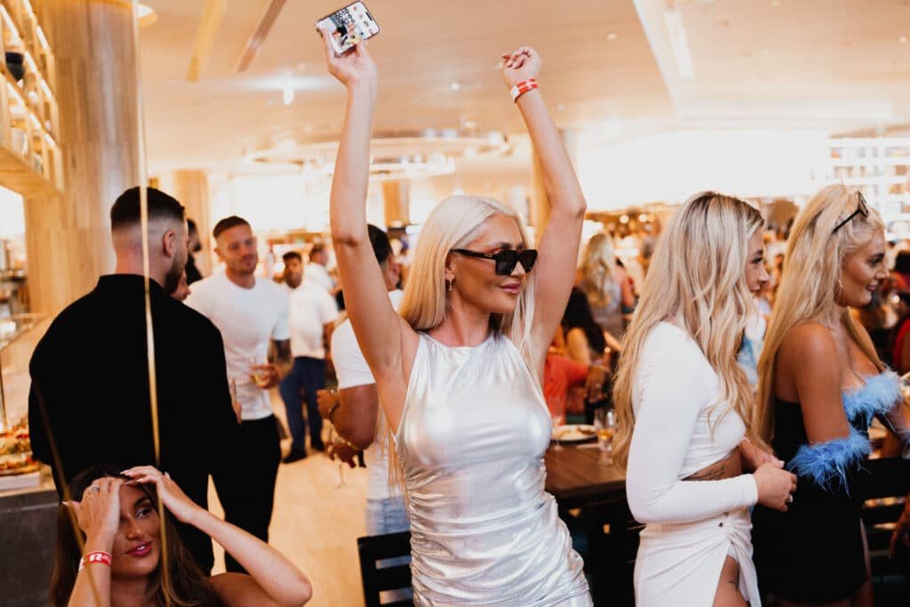 A woman in a silver dress raises her phone in the air, surrounded by people in The Best Fun Brunches in Dubai setting.