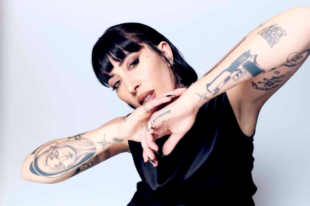 A person with fringe bangs shows off arm tattoos, displaying a confident pose against a plain background.