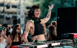 A DJ with headphones around their neck is energetically mixing music at an outdoor event, surrounded by a crowd.