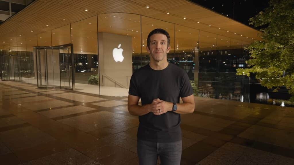 A person stands outside a glass-fronted Apple store at night, wearing a black shirt and watch.