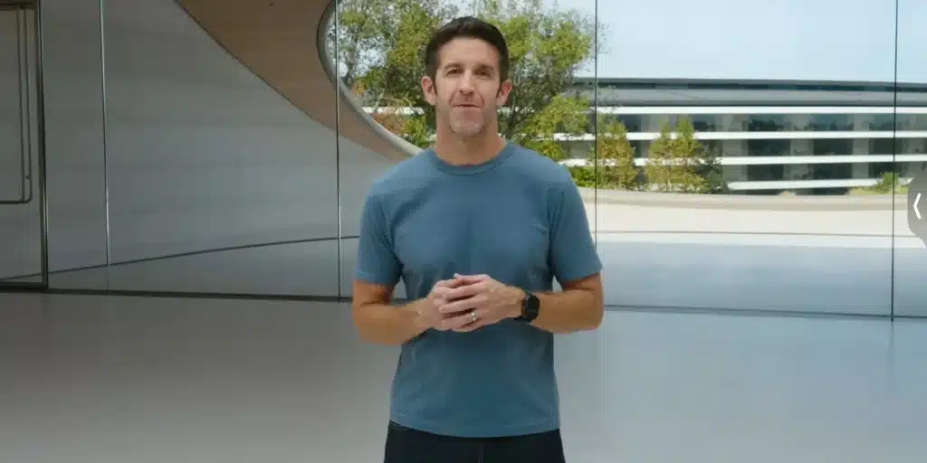 John Ternus in a blue shirt stands in a modern, open space with large windows and greenery outside.