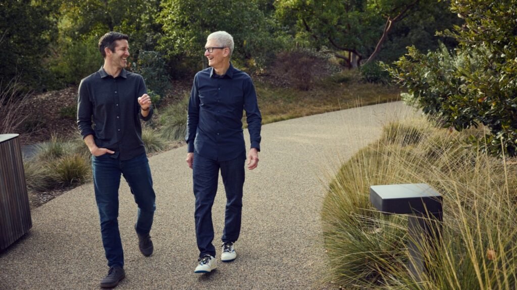 John Ternus and Tim Cook are walking on a gravel path through a lush, green park, engaged in conversation.
