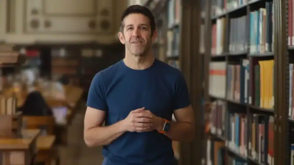 A person in a blue shirt stands in a library with shelves of books and others studying in the background.