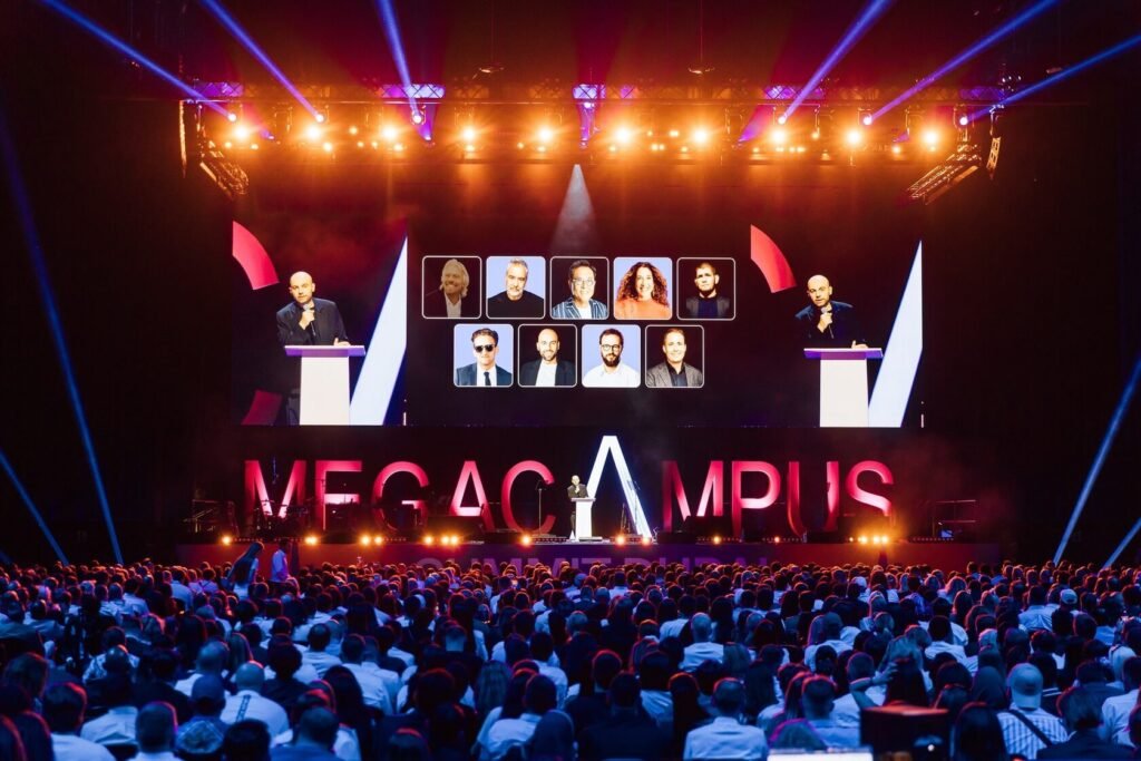 A speaker stands on stage with "MEGACAMPUS" displayed, large crowd in attendance, and various portraits projected above.