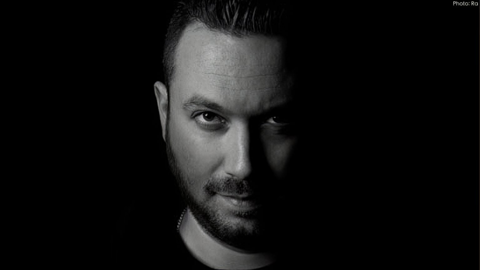 Man with short hair and beard illuminated on one side, set against a dark background.