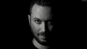 Man with short hair and beard illuminated on one side, set against a dark background.