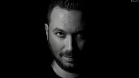 Man with short hair and beard illuminated on one side, set against a dark background.