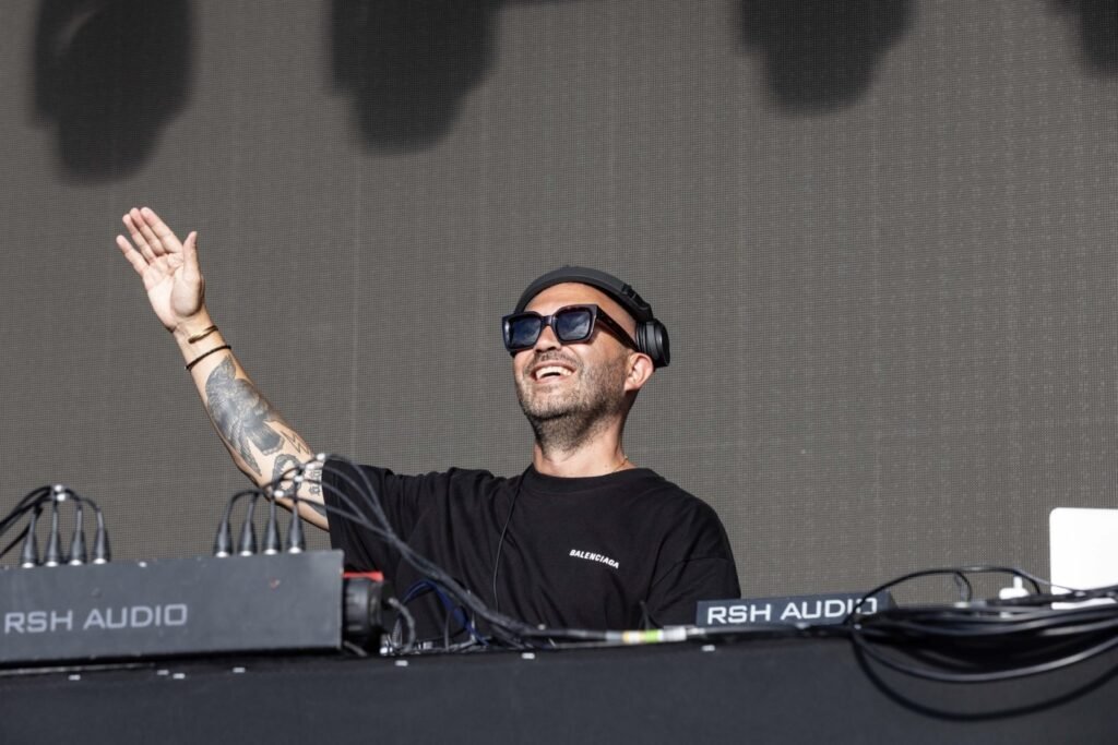 Nic Fanciulli, wearing sunglasses and a black shirt is performing on stage, smiling with one hand raised.