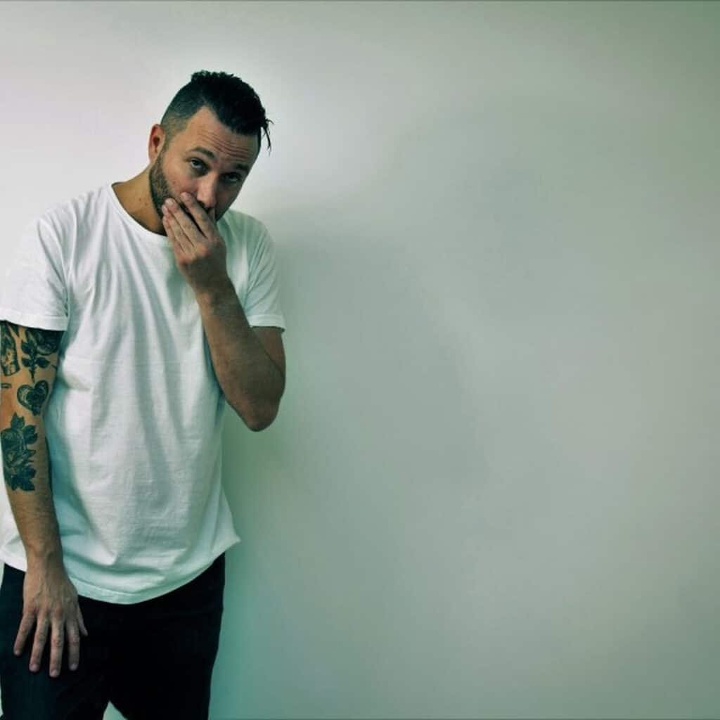 Nic Fanciulli with tattoos on his left arm, wearing a white T-shirt and black pants, stands against a plain background, covering his mouth.