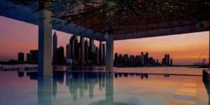 A stunning city skyline at sunset reflects on a calm pool, with a modern roof structure above.
