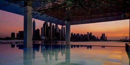 A stunning city skyline at sunset reflects on a calm pool, with a modern roof structure above.