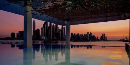 A stunning city skyline at sunset reflects on a calm pool, with a modern roof structure above.